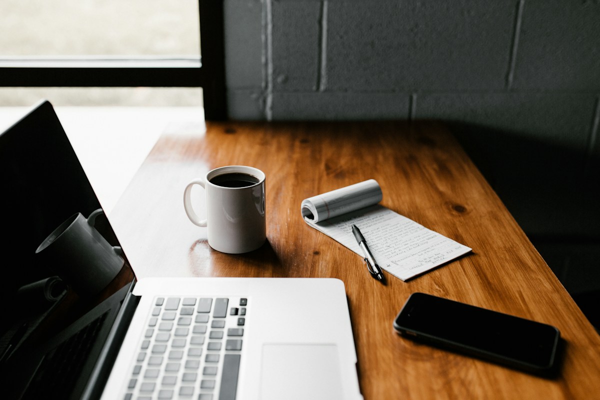 An open notebook, coffee and laptop on a sunlit desk